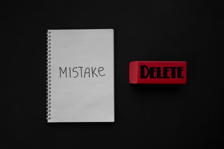A white spiral-bound notepad with the word 'MISTAKE' handwritten in black ink is positioned on a black background. Next to it, there is a red rubber stamp with the word 'DELETE' in bold black letters.